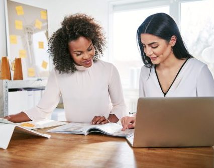 Two young women working together in an office at their small business sitting reading a report or paperwork with pleased smiles